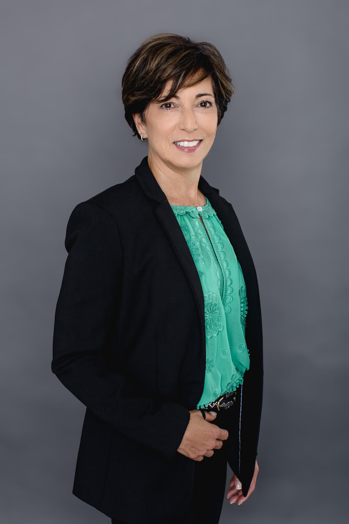 A woman with black hair standing in front of a gray backdrop wearing a black suit and a green shirt with one hand in her pocket smiling at the camera