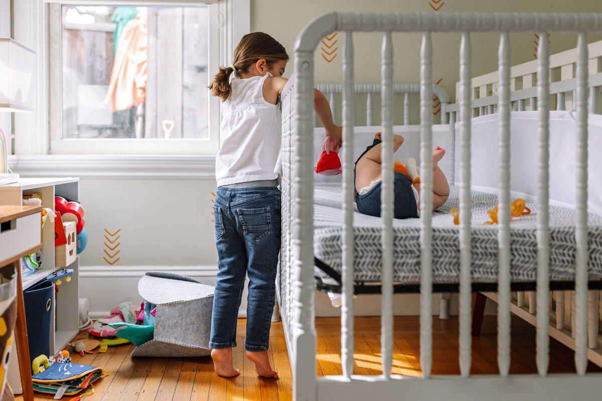 sister reaching on tiptoes into crib to interact with baby sister who has feet up