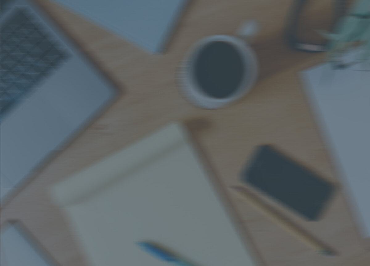 blurred image in background of graphic designers desk with laptop, coffee mug, notepad, phone and pencil