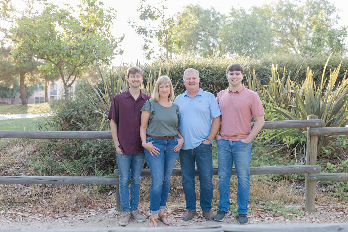 A family of four standing in front of a wooden park fence and lots of green trees and bushes, smiling at the camera with their hands in their jean pockets. Captured at Citrus Ranch Park in Tustin.