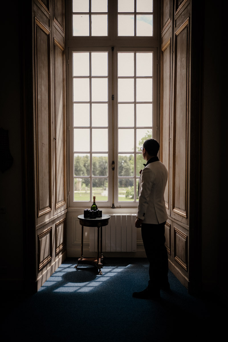 Le marié asiatique termine ses préparatifs dans une pièce baignée de lumière au Château de Saint-Martin-du-Tertre, juste avant la cérémonie de mariage.