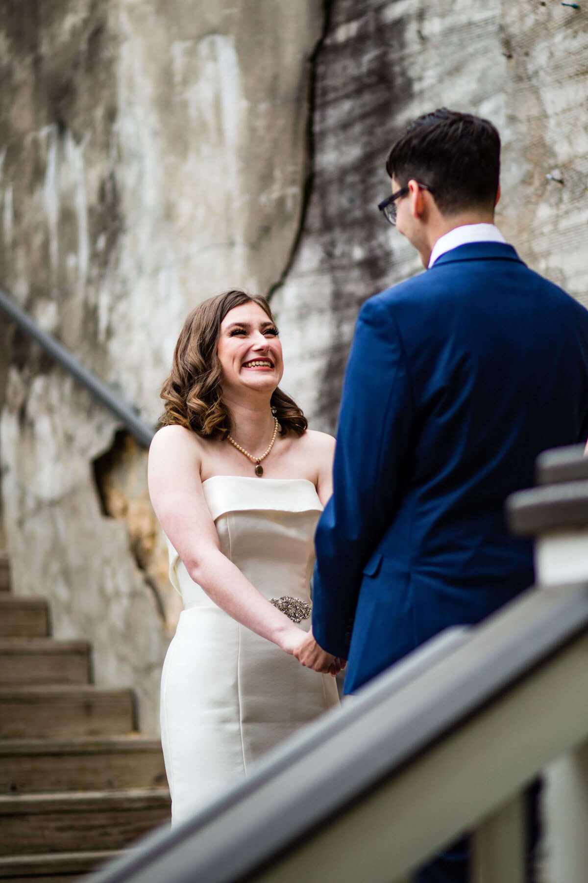 A bride in a white strapless dress smiles joyfully at her groom, who is wearing a blue suit, as they hold hands on wooden stairs next to a textured, weathered wall.