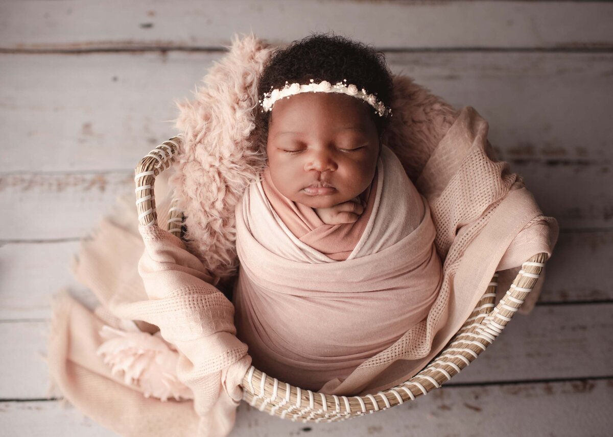 Aerial image of baby girl sleeping in basket with fabric draping over the edges. Baby is wearing a blush wrap  and delicate floral headband.