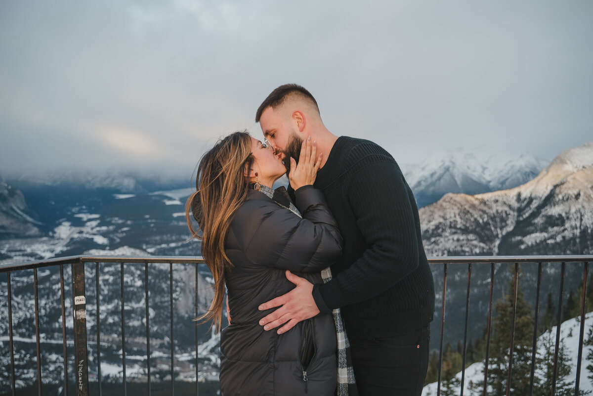 Couple kisses with mountains behind during snowy Sulphur Mountain proposal.