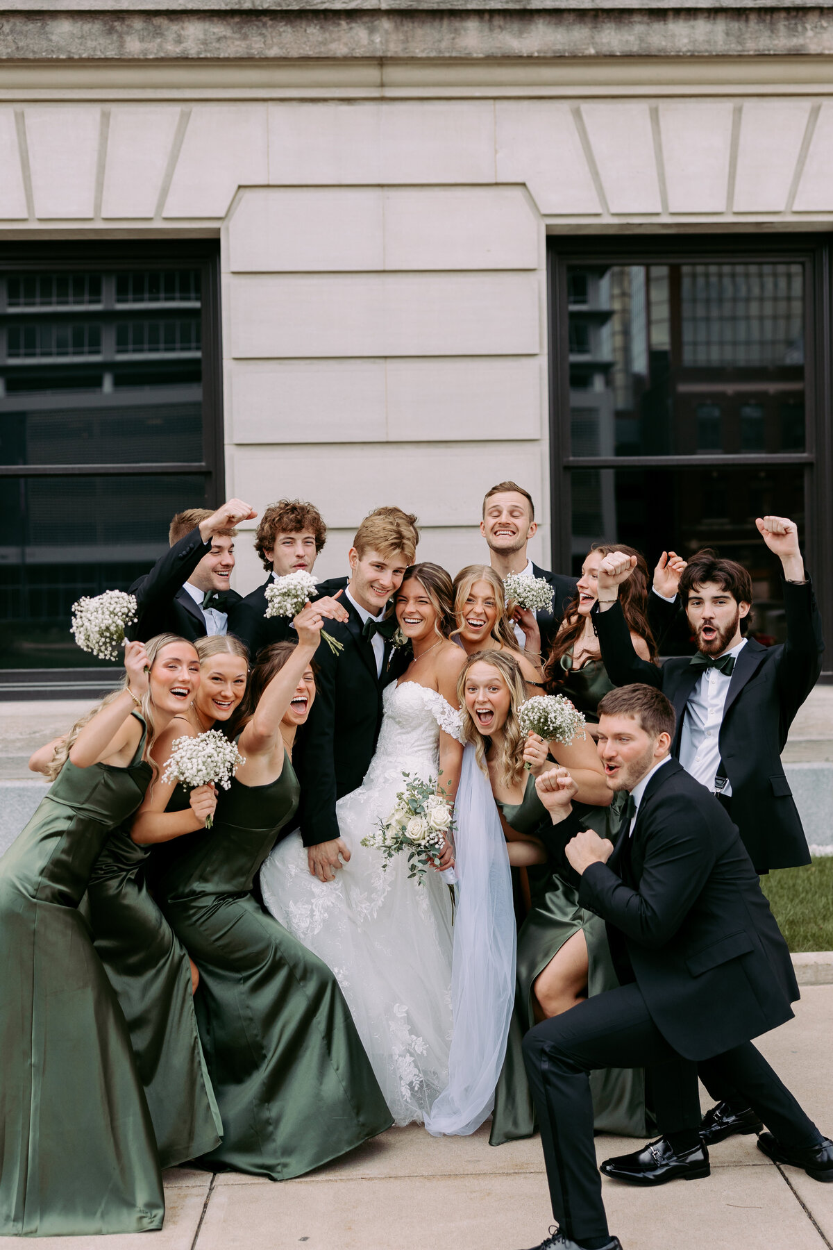 This photo shows a bridal party all piled around the bride and groom on a wedding day. This photo is in front of the Allen County Courthouse in Fort Wayne, Indiana.