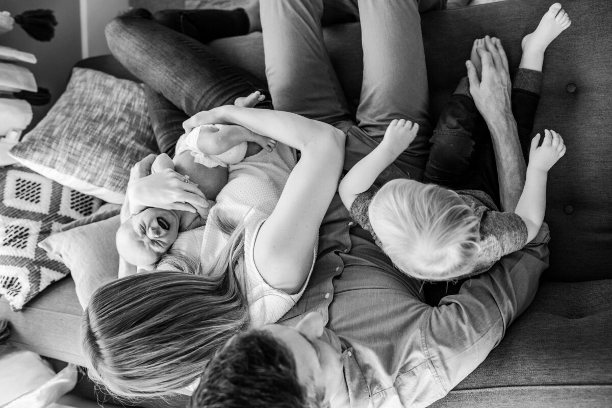 Black and white image of a family sitting on a couch. The mother holds a crying newborn, while a toddler sits on his father's lap, with everyone's legs intertwined.