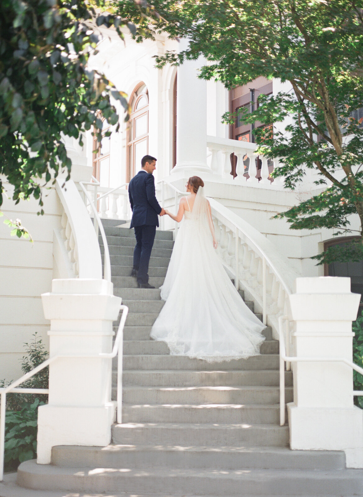 Bride and groom on stunning white elegant staircase, bridal gown with flowing train, captured by Ana Douglas Photography, timeless and authentic wedding photographer in Vancouver, BC. Featured on the Bronte Bride Vendor Guide.