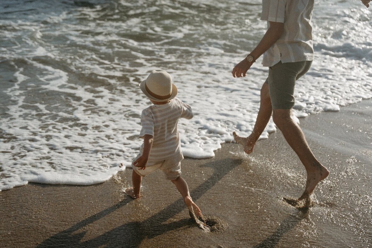 Le père et son fils courent pieds nus sur le sable mouillé par l'écume. L'enfant porte un chapeau en paille. Séance photo famille à la plage par Kamescop.
