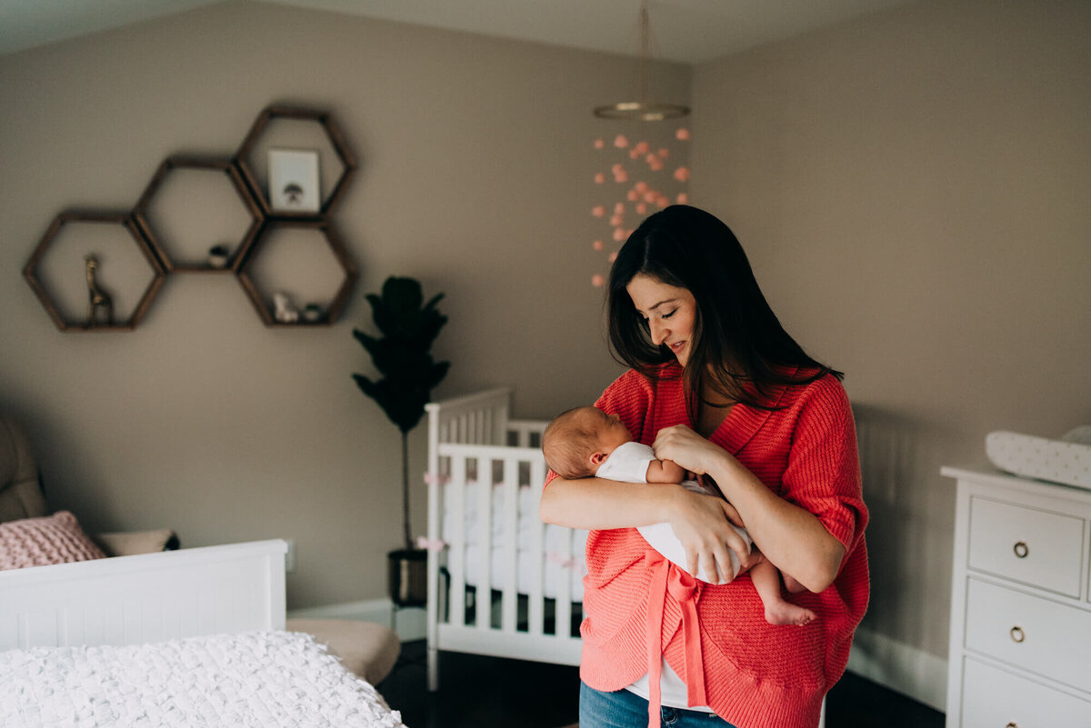 A mother wearing a pink sweater holds her daughter in her nursery that has white and pink accents