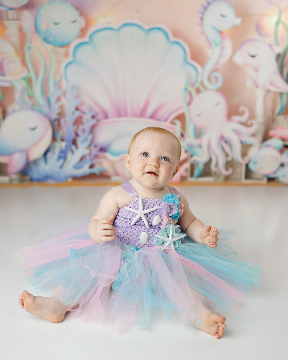 Baby in a pastel mermaid tutu sitting in front of an under-the-sea backdrop during a cake smash session. Photograph by Yucaipa cake smash photographer Kaitlyn Dawn Photography.