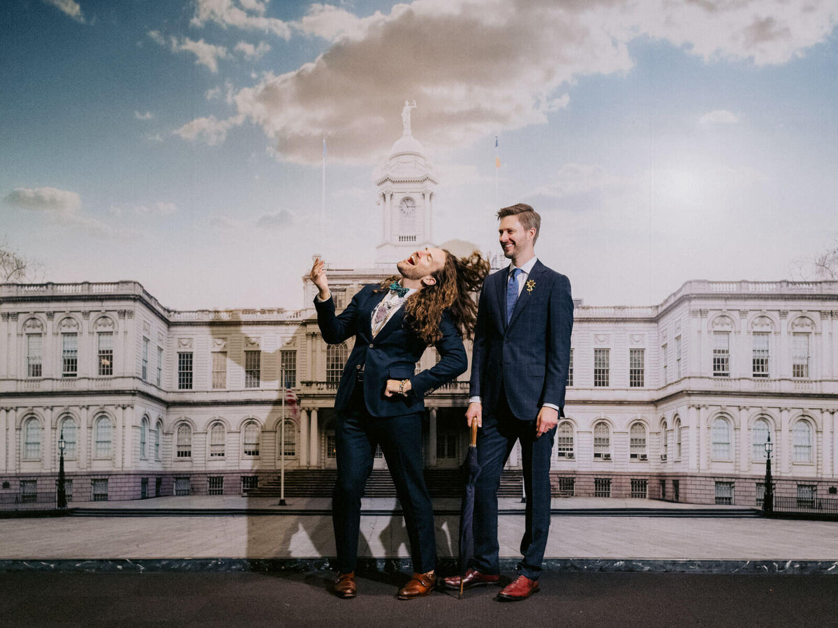 The groom is doing a wacky shot as the other groom watch on, in front of the NYC City Hall wall photo. Elopement Image by Jenny Fu Studio