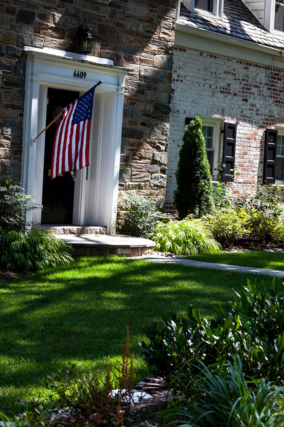 Stone and brick house with an American flag by the front door, surrounded by a lush green lawn and landscaped garden