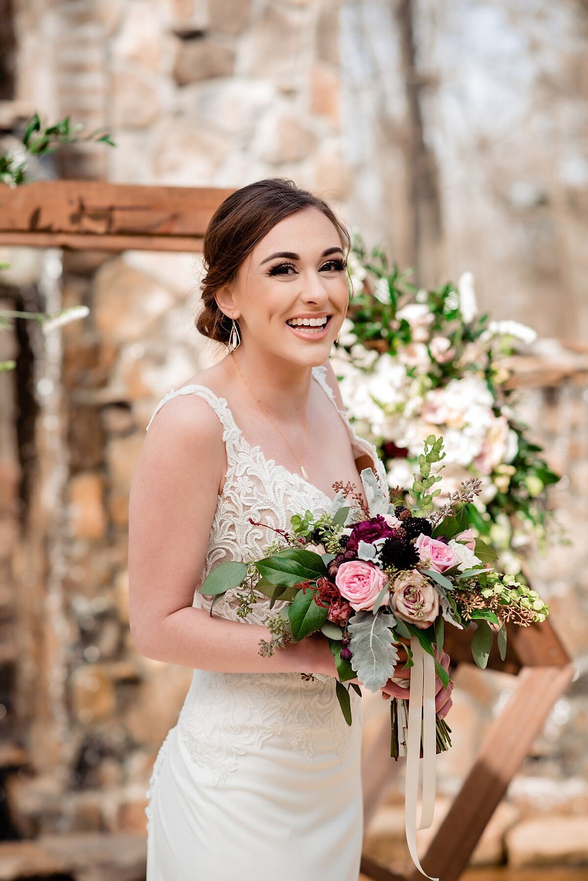 The bride stands at the wooden arbor in a lace, fitted wedding dress with lace straps. She is holding a horizontal bouquet of burgundy and blush roses and greenery accented by long sheer ribbons. The arbor behind them has a large spray of blush and ivory flowers with greenery.
