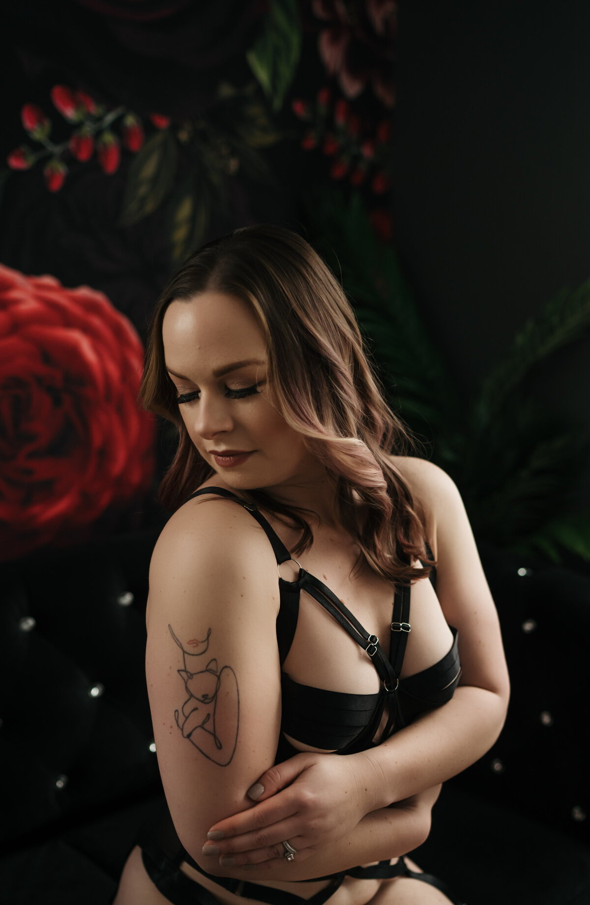 A woman in black strap lingerie gazes down her shoulder while sitting on a black couch in front of a rose tapestry in a studio