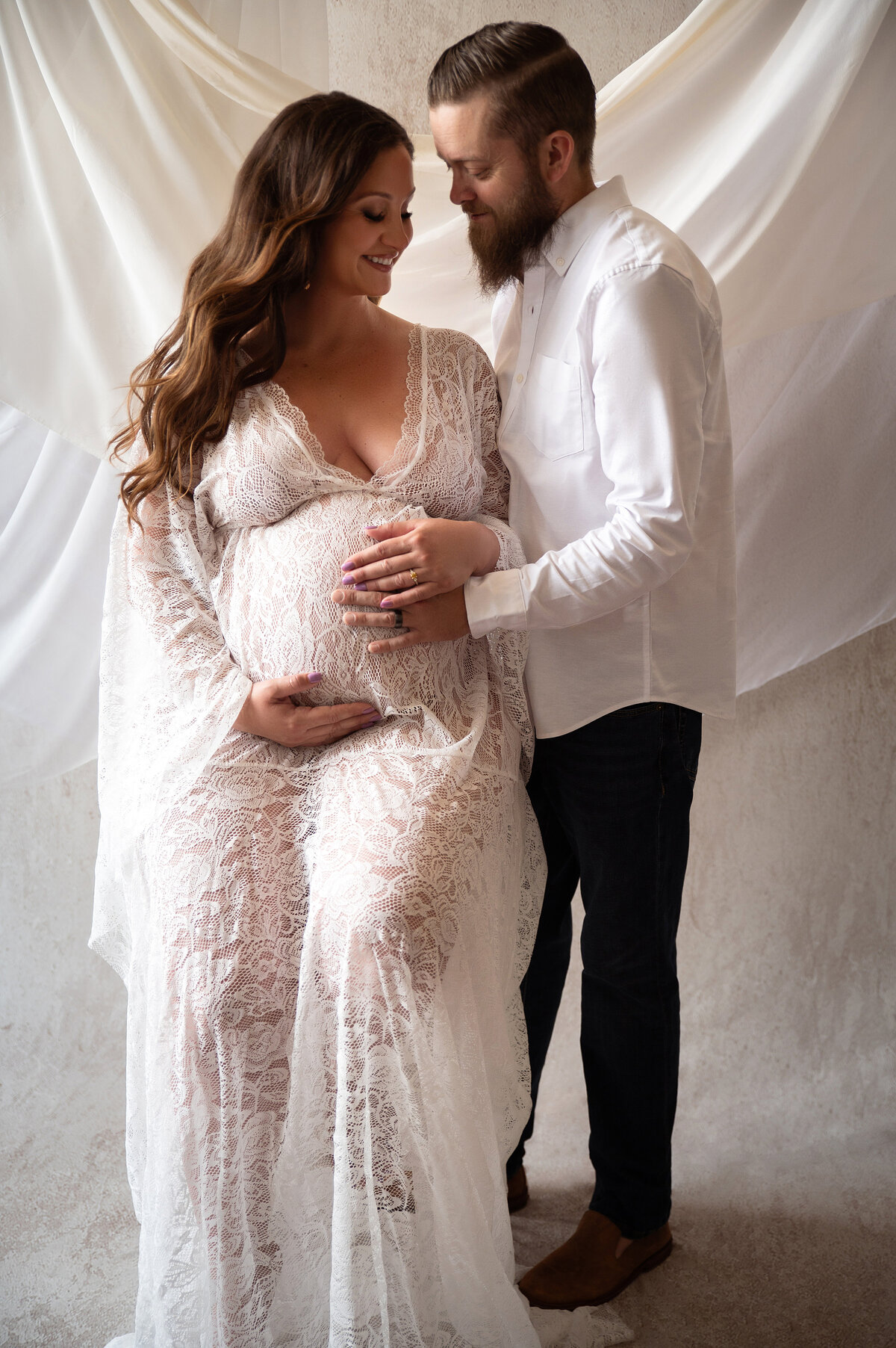 Parents to be excitedly hold mom to be's baby bump while looking down at mom's belly wearing white in front of white drapes background