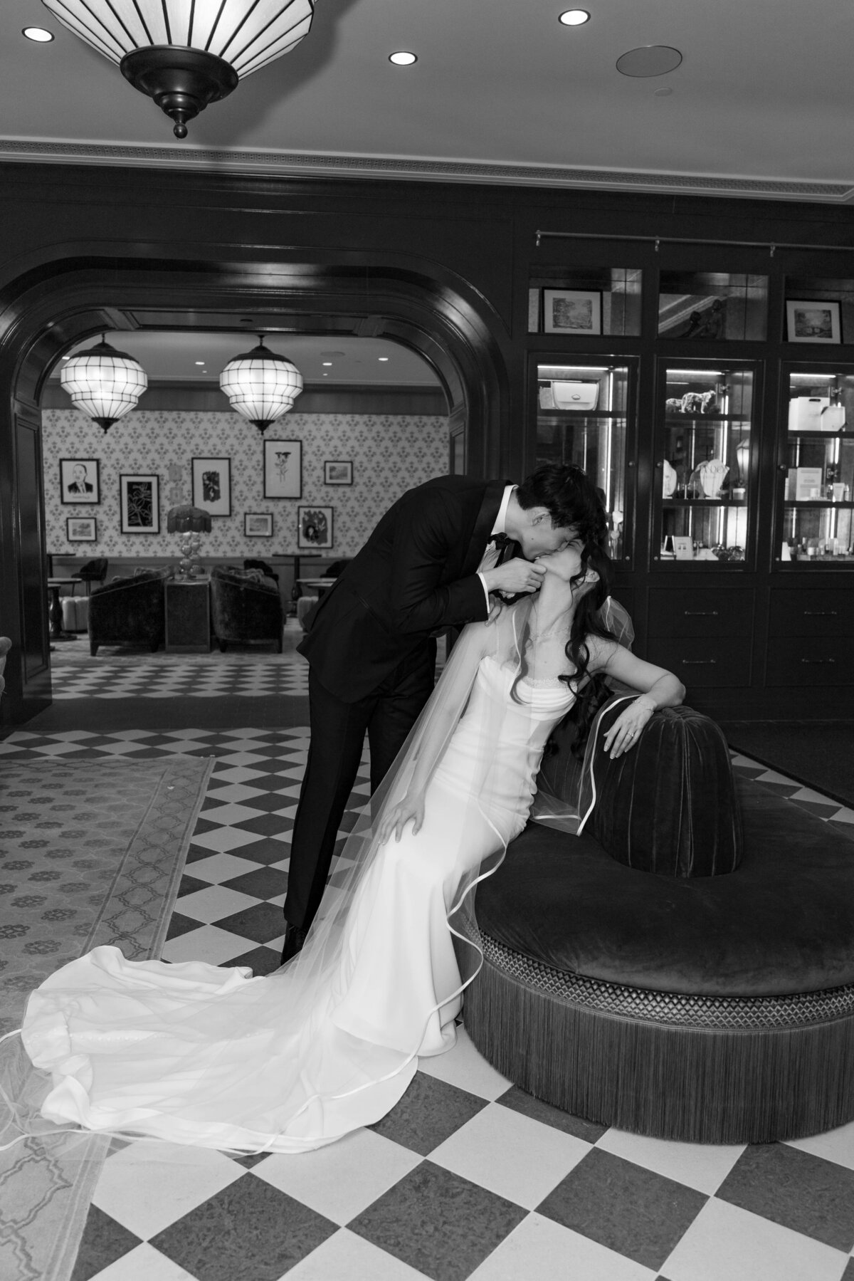 Rhode Island Wedding Photographers | Kelsey Sheehan Photography Timeless Rhode Island Weddings | A bride in a flowing gown sits on a sofa, leaning back as a groom in a suit tenderly kisses her. The setting is an elegantly decorated room.