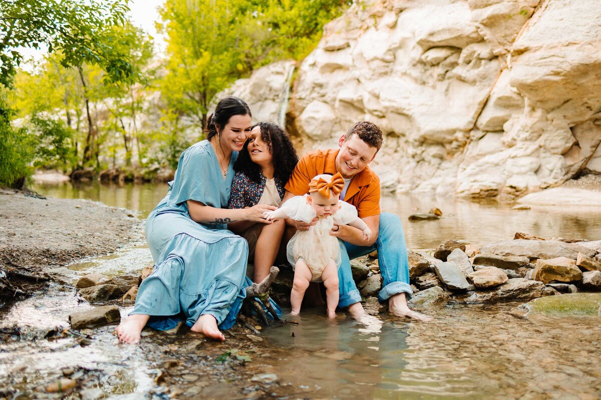An incredible family plays with their baby by a tranquil lake and lush forest. Captured in a joyful moment, they interact with the water and enjoy their time together in a beautiful natural setting.