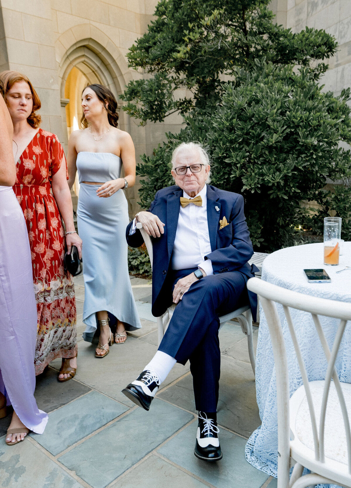 The photo was taken at Washington D.C., specifically at the Washington National Cathedral by the talented photographer Twah Dougherty. The setting captures the serene beauty of the cathedral grounds. In the foreground, an elderly man dressed in a suit is seated at a table, his stylish appearance enhanced by the addition of glasses and eye-catching black-and-white shoes. Flanking him are two women, one elegantly dressed in a striking red and white ensemble and the other in a charming light blue off-the-shoulder dress. The scene is beautifully framed by lush trees and the imposing stone walls of the cathedral in the background. Additionally, the current films as videographers also captured this moment, adding a dynamic visual element to the overall ambiance of the photograph.