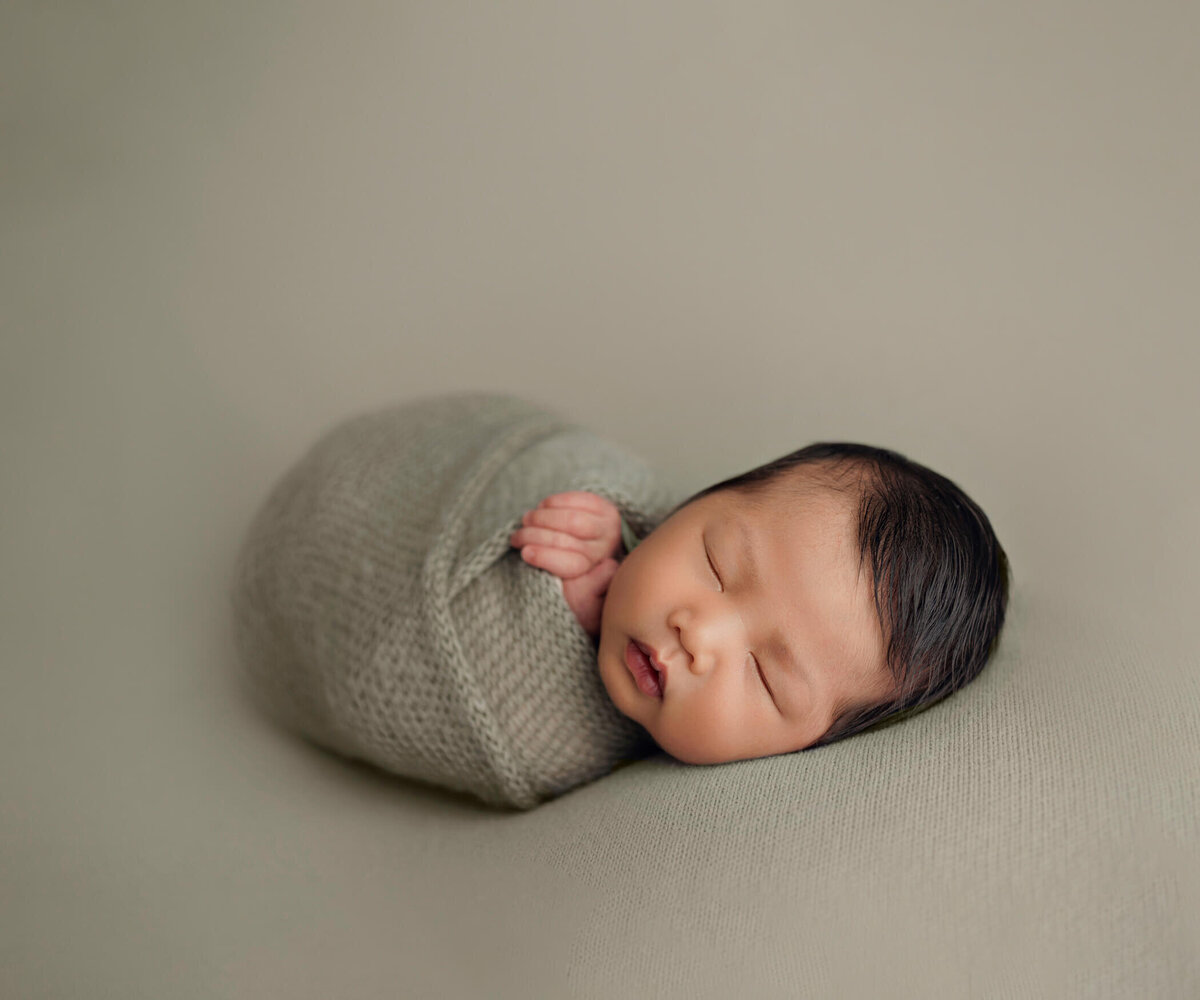 A peaceful newborn baby sleeps soundly, swaddled in a soft, light green wrap against a matching light green background. The baby's serene expression and gentle pose evoke a sense of calm and tranquility.