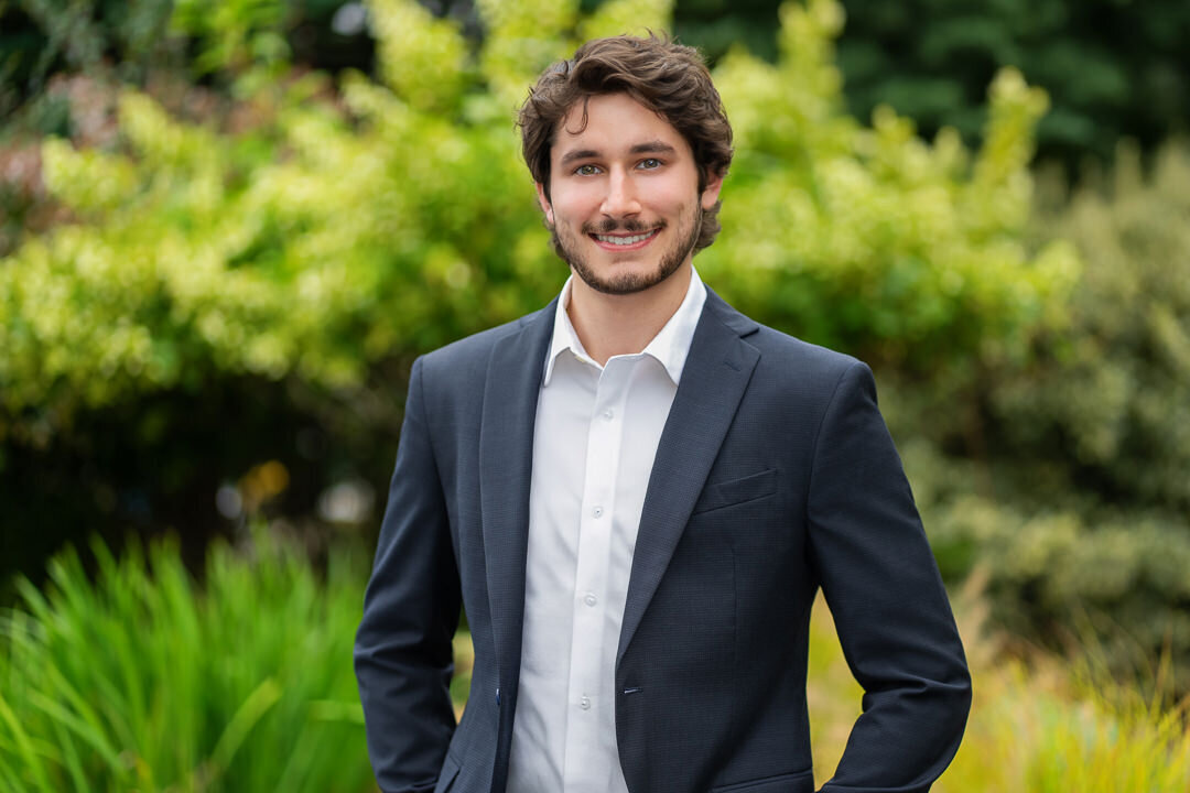 Outdoor headshot of smiling young man in navy blazer and white shirt with lush green background – NJ business headshot photographer