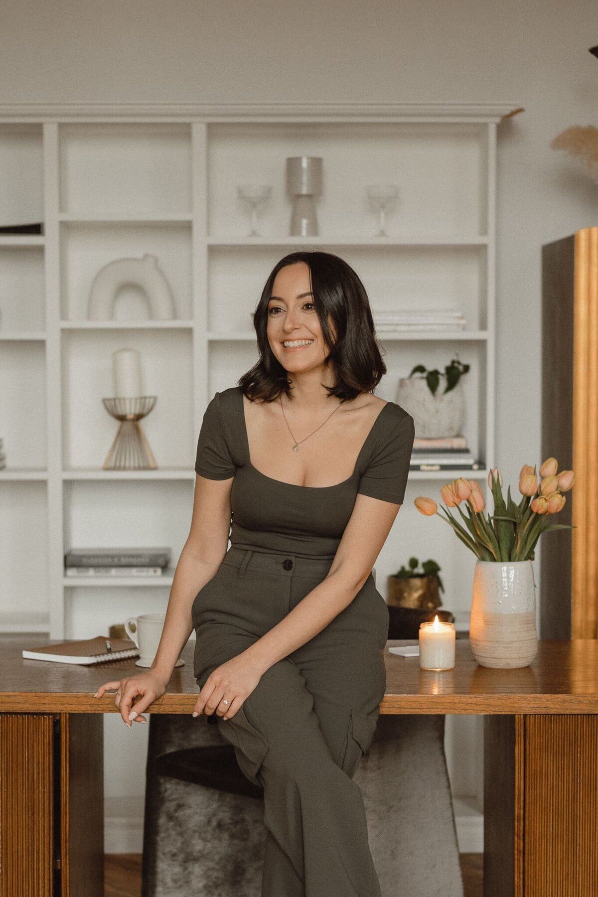 Vickie of VERVE & COLOR leaning on a table with a lit candle and tulips, looking away and smiling happily, embodying a chic and professional vibe.