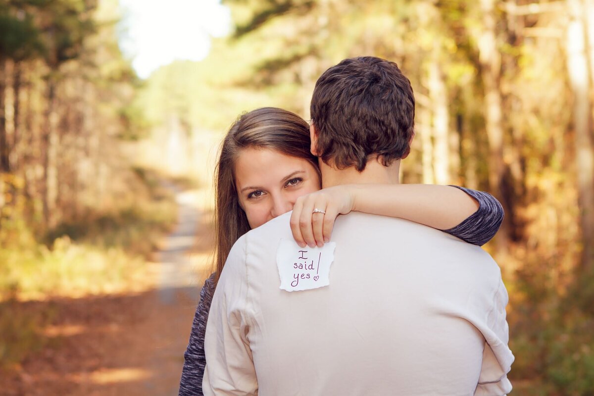 Fun engagement portrait of a women looking over the shoulder of her fiance holding a note that says I said yes.