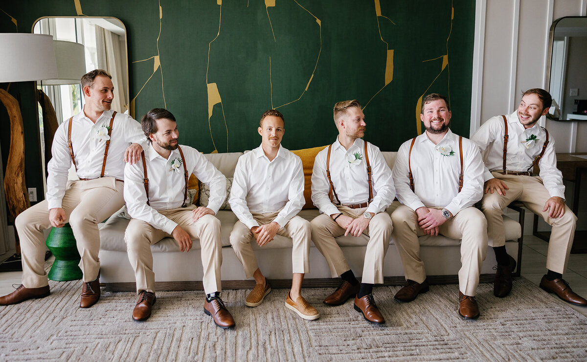 Hunter and his groomsmen, dressed in beige suits and suspenders, sit in a stylish indoor space before the ceremony.