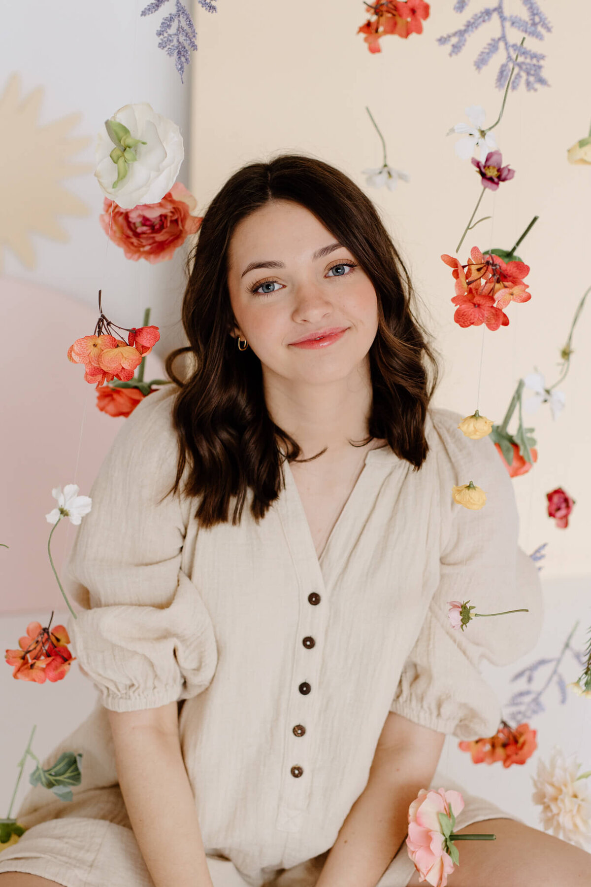 Senior portrait of 2023 graduate in off white romper sitting in front of floral wall in photography studio