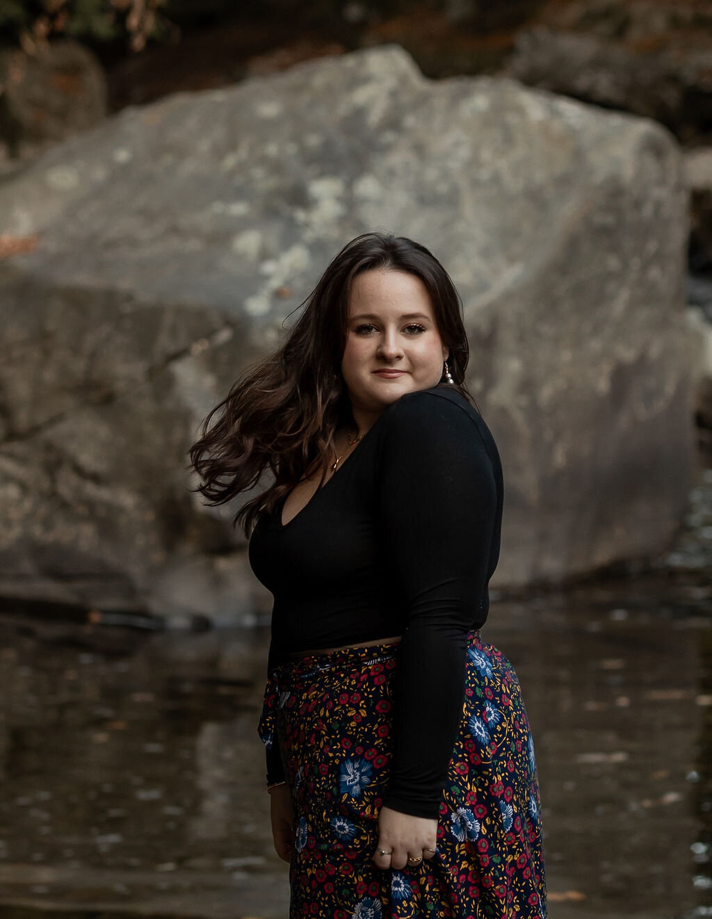 A person with long dark hair, wearing a black long-sleeve top and a floral skirt, stands near water with a large rock in the background. Capturing this serene moment, a New England Portrait Photographer perfectly frames their calm expression against nature's backdrop.