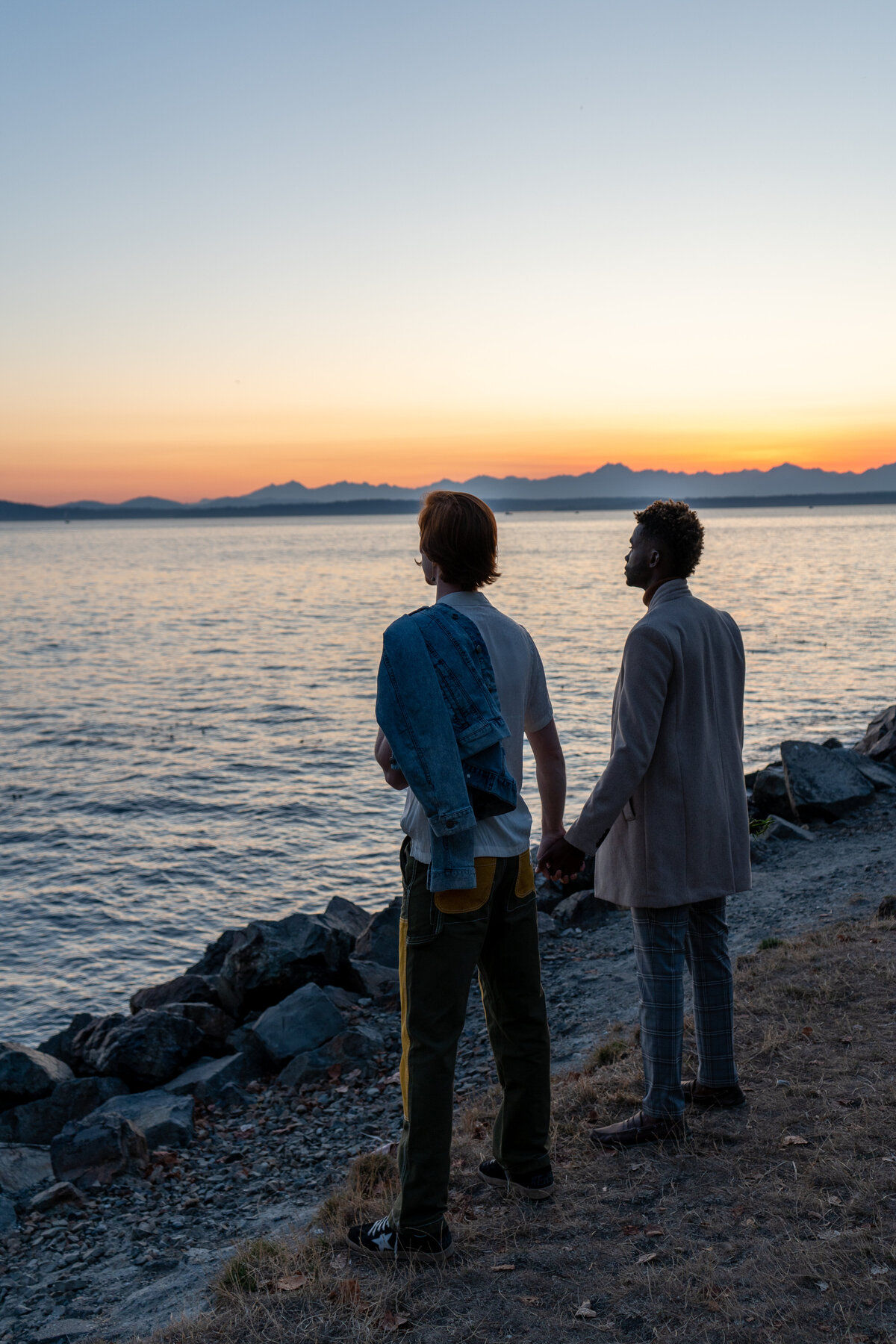 Gay couple at Pocket Beach, Seattle at sunset looking out at the ocean while holding hands