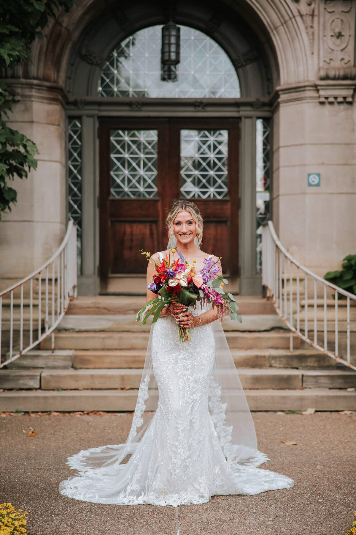 A bride in an elegant lace gown holding a vibrant bouquet stands before the historic entrance of The Phipps Conservatory.