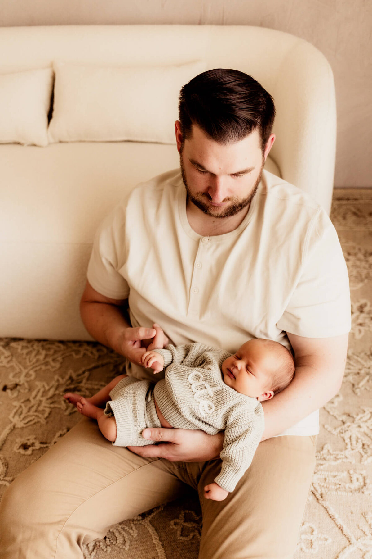 Father looks down at his son while holding him and sitting on a beige rug.