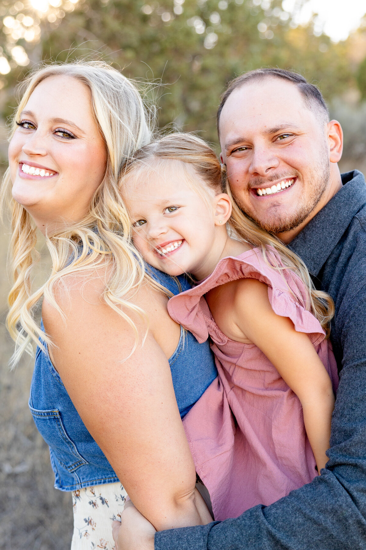 A mother, father, and their daughter lean into each other, grinning and embracing, with sunlit trees and desert shrubs in the background.