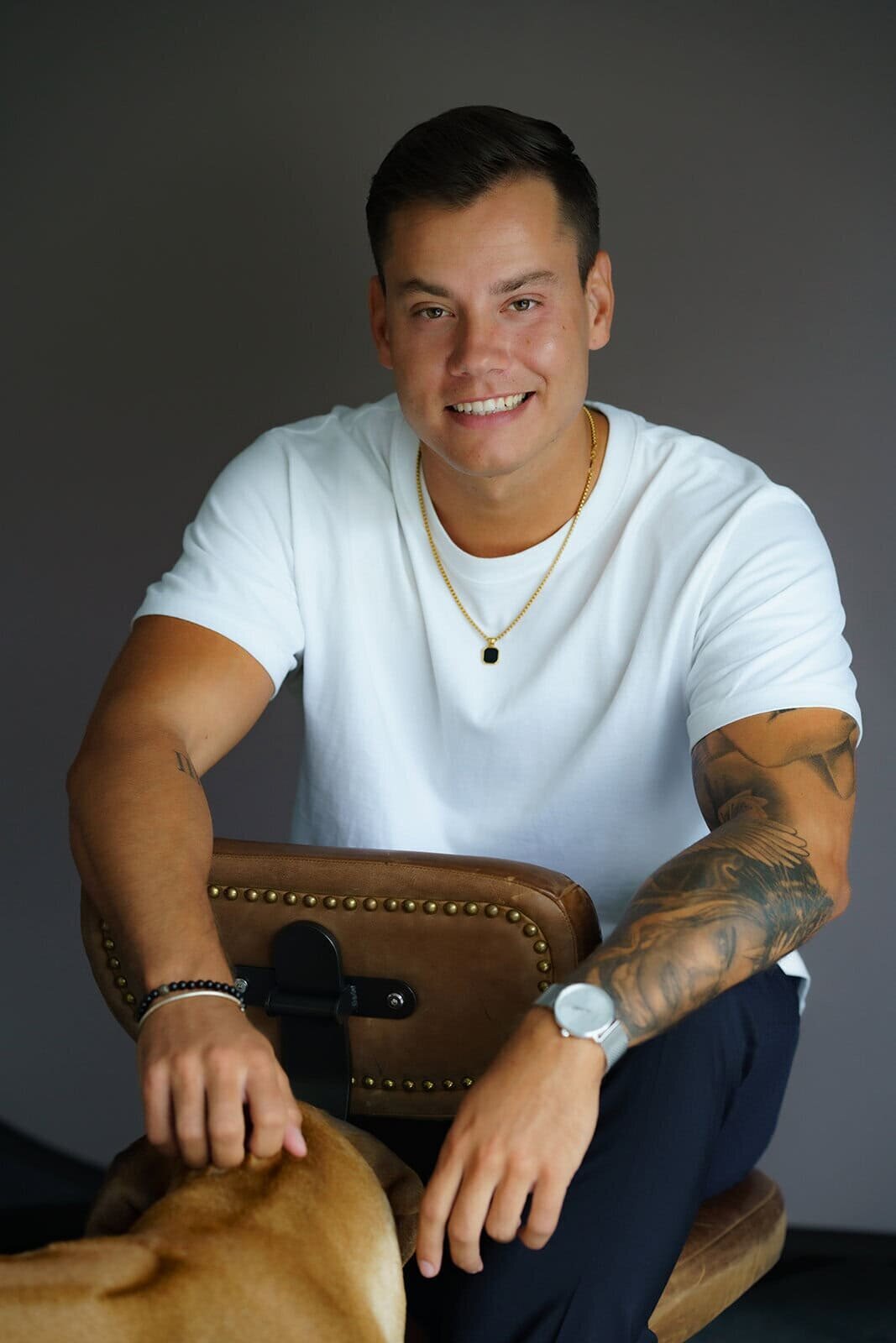 Young man sitting backwards in a small chair smiling and wearing a white t-shirt and a small necklace with large tattoos on his arm.