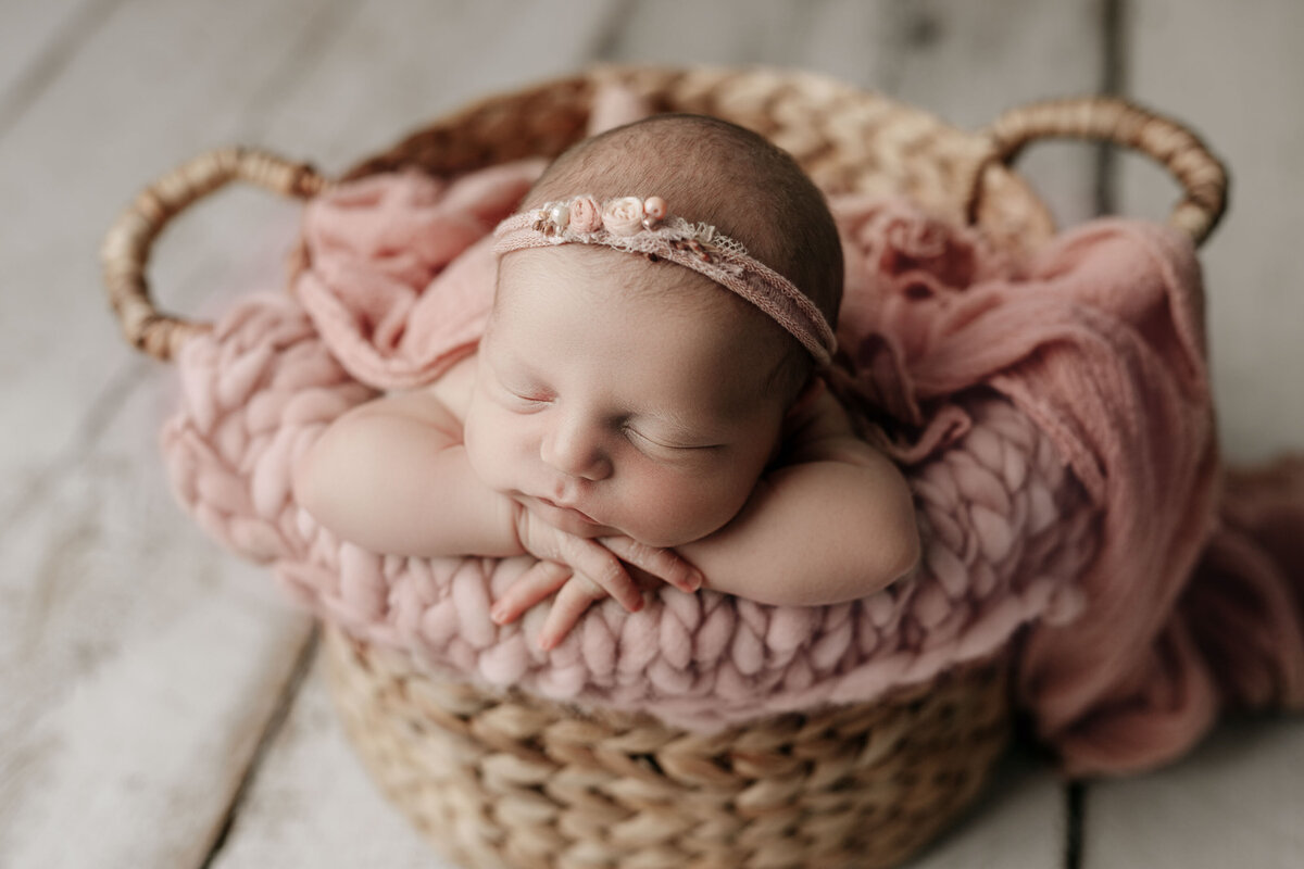 Baby sleeping in woven basket with a dusty pink swaddle flowing over the edges. Baby is resting her chin on folded hands and wearing a delicate floral headband.