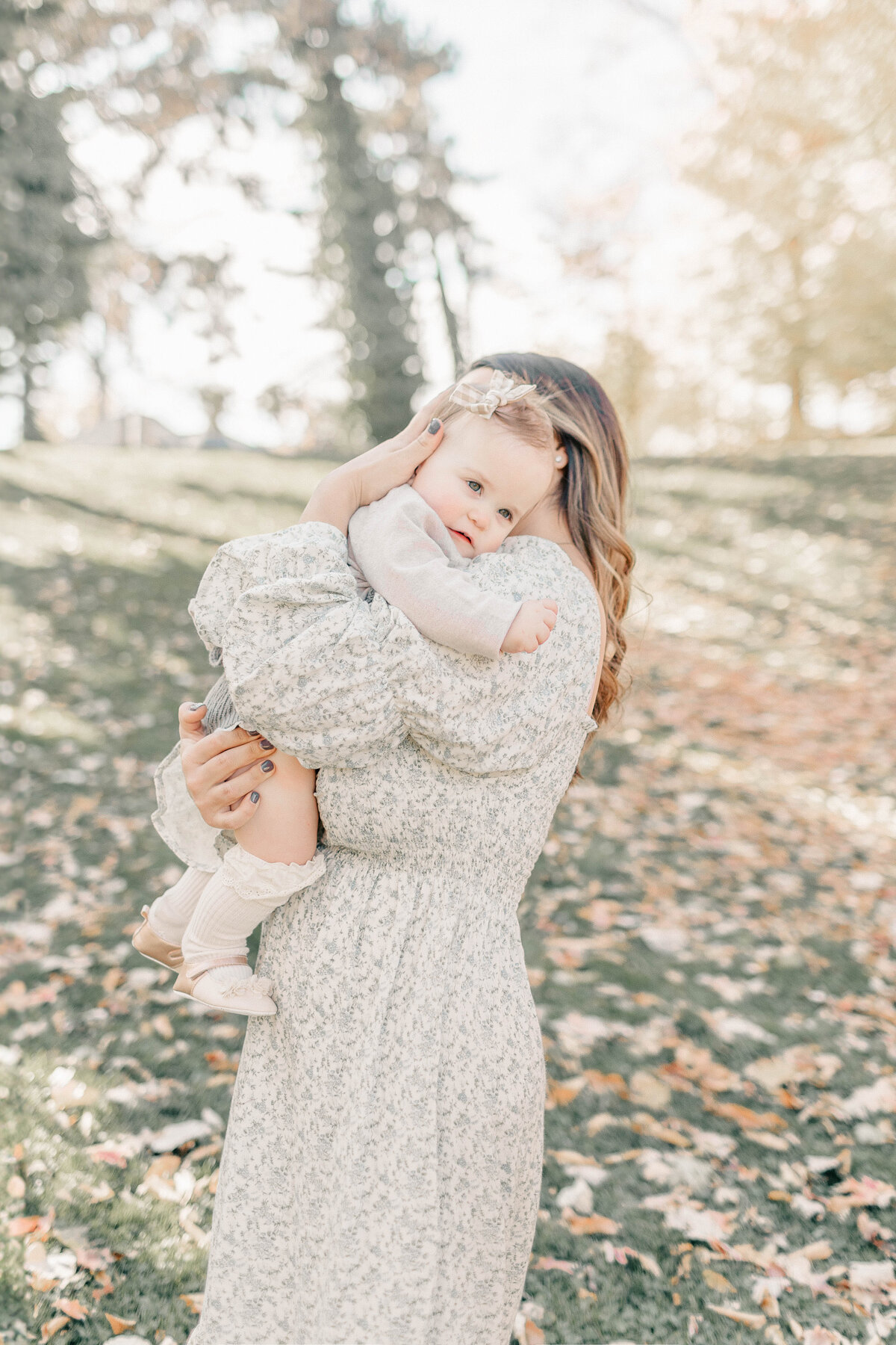 A mother wearing a floral dress snuggles her daughter during her first birthday milestone session at at Kansas City park. 