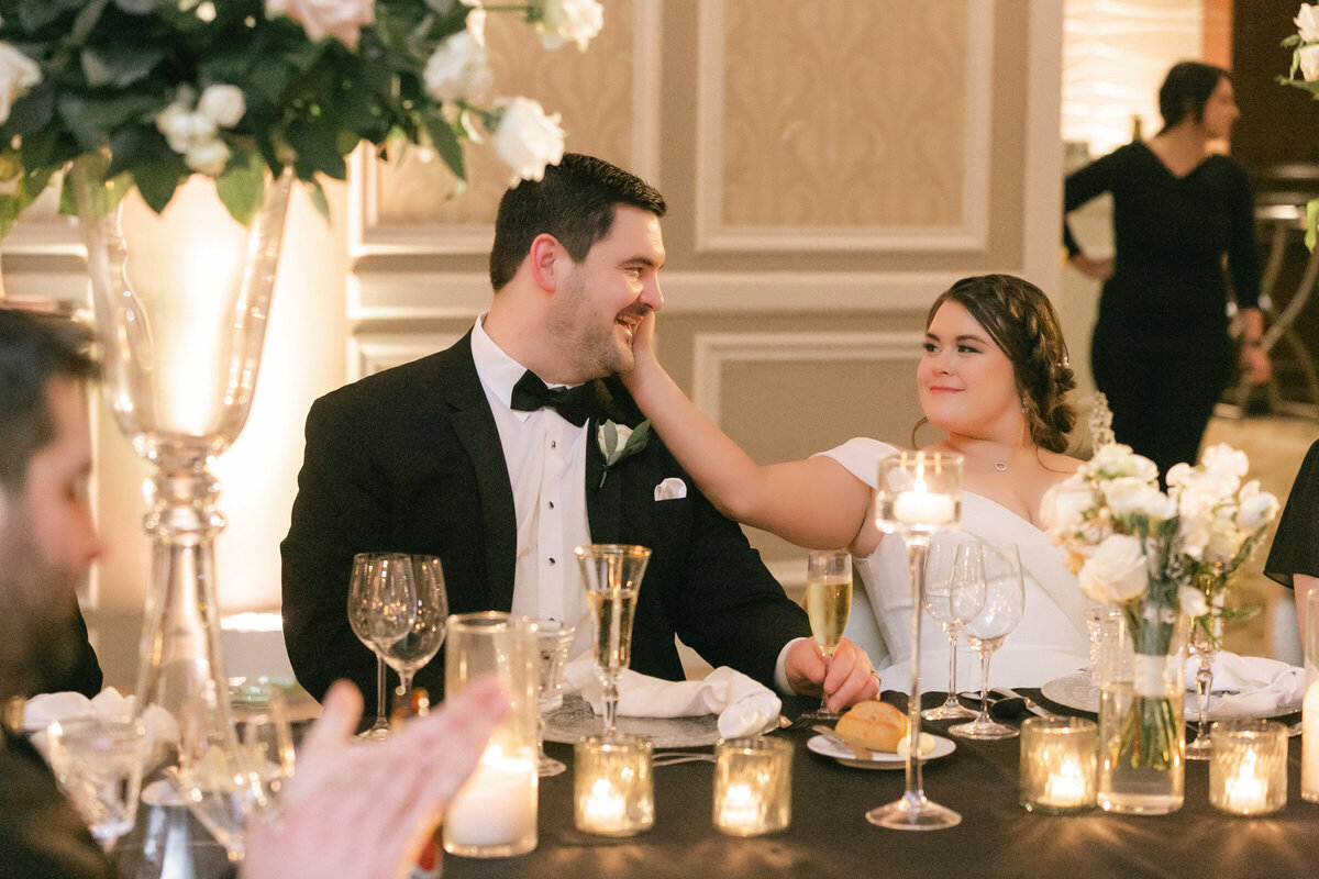 bride and groom at their reception table at The Adolphus in Dallas, sharing a tender moment as she gently touches his cheek, smiling at each other with love in their eyes.