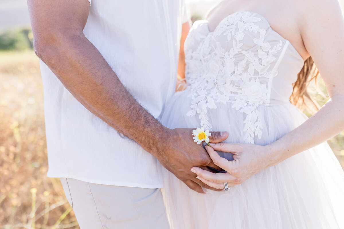 A couple standing in a sunny field, with the womans hand gently resting on her pregnant belly, holding a small daisy flower. Both are wearing light-colored clothing, and the focus is on the intricate lace details of her dress.