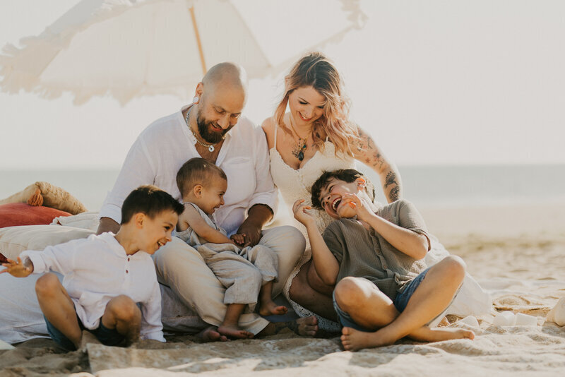 Deux parents et leurs trois enfants assis sous une ombrelle riant ensemble pendant un shooting photo en Vendée, à la plage.