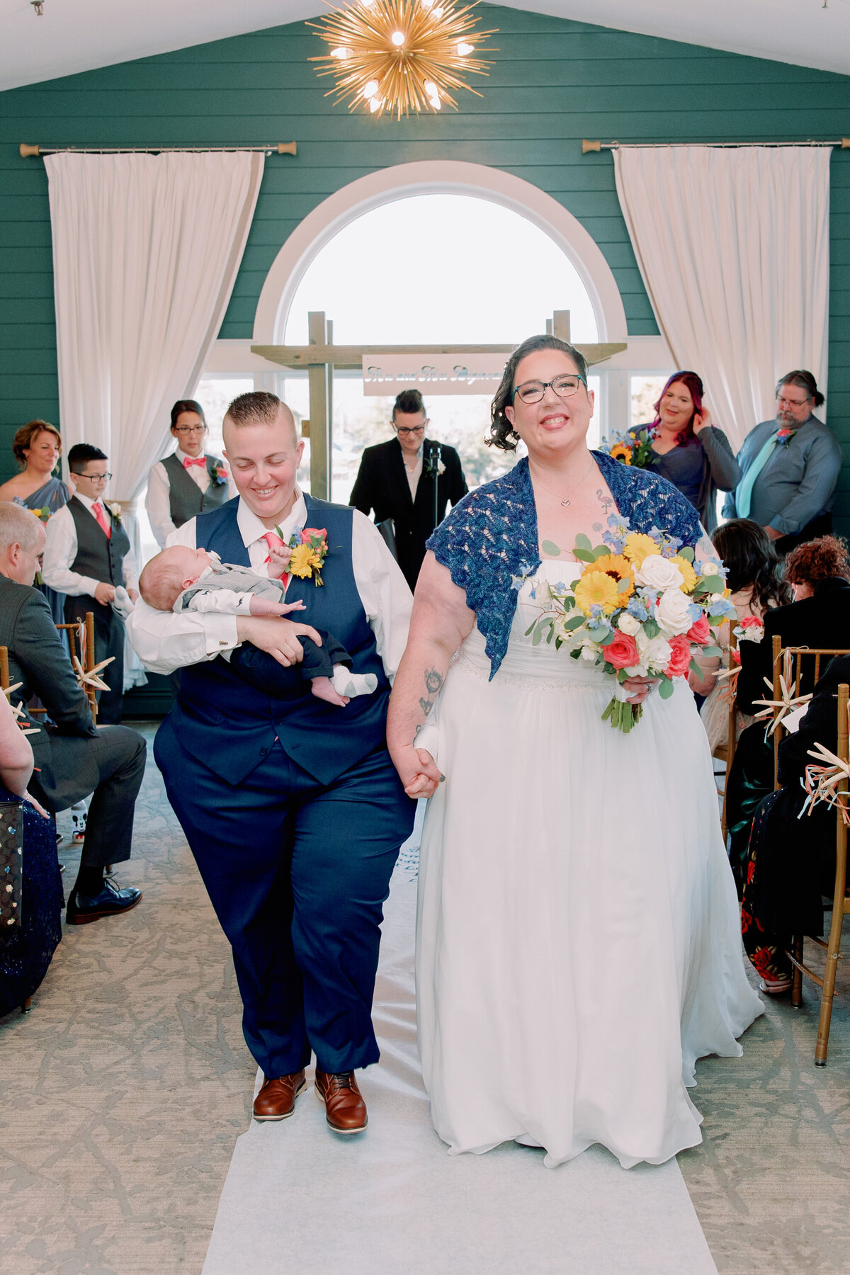 A newlywed couple holding hands and walking back up the aisle as one also holds a newborn 