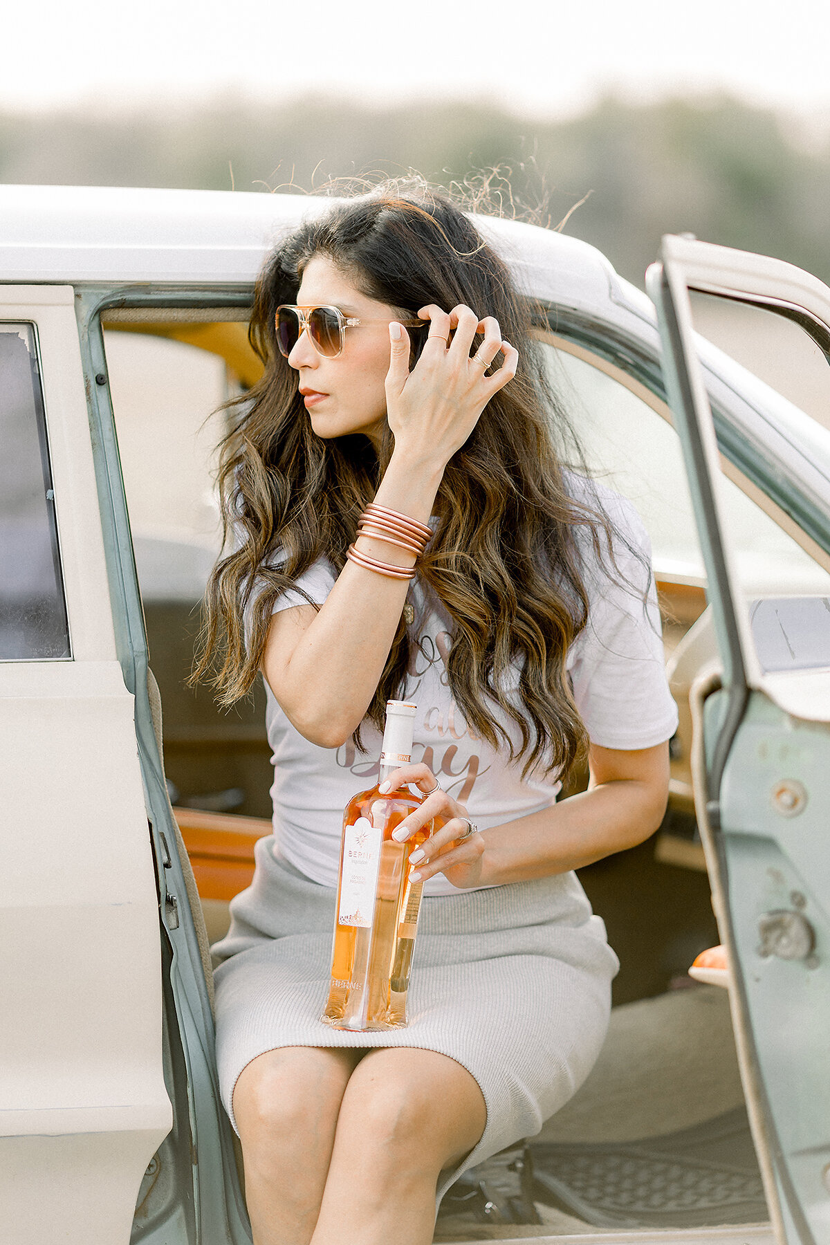 Professional headshot photo taken at a Dallas/Fort Worth lake of a local business owner sitting in a vintage car while she is holding a bottle of wine and holding her hair back from the wind.