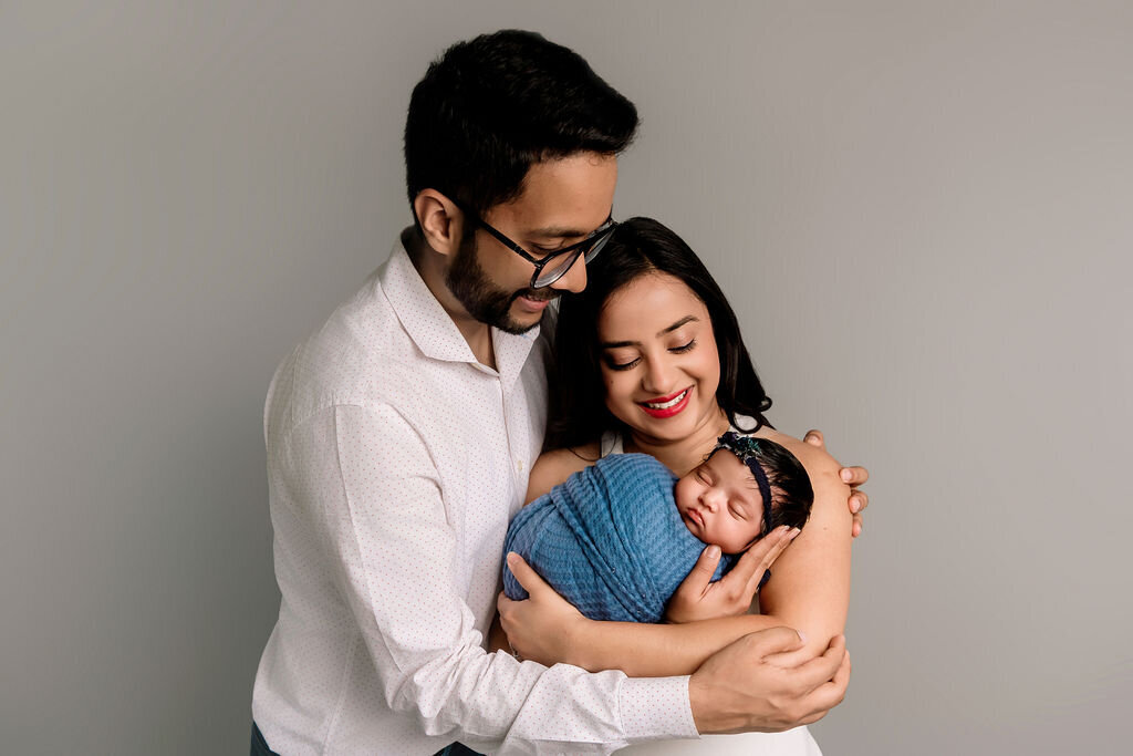 Connecticut newborn photographer captures tender family portrait with new parents embracing their sleeping baby wrapped in blue blanket against neutral studio backdrop