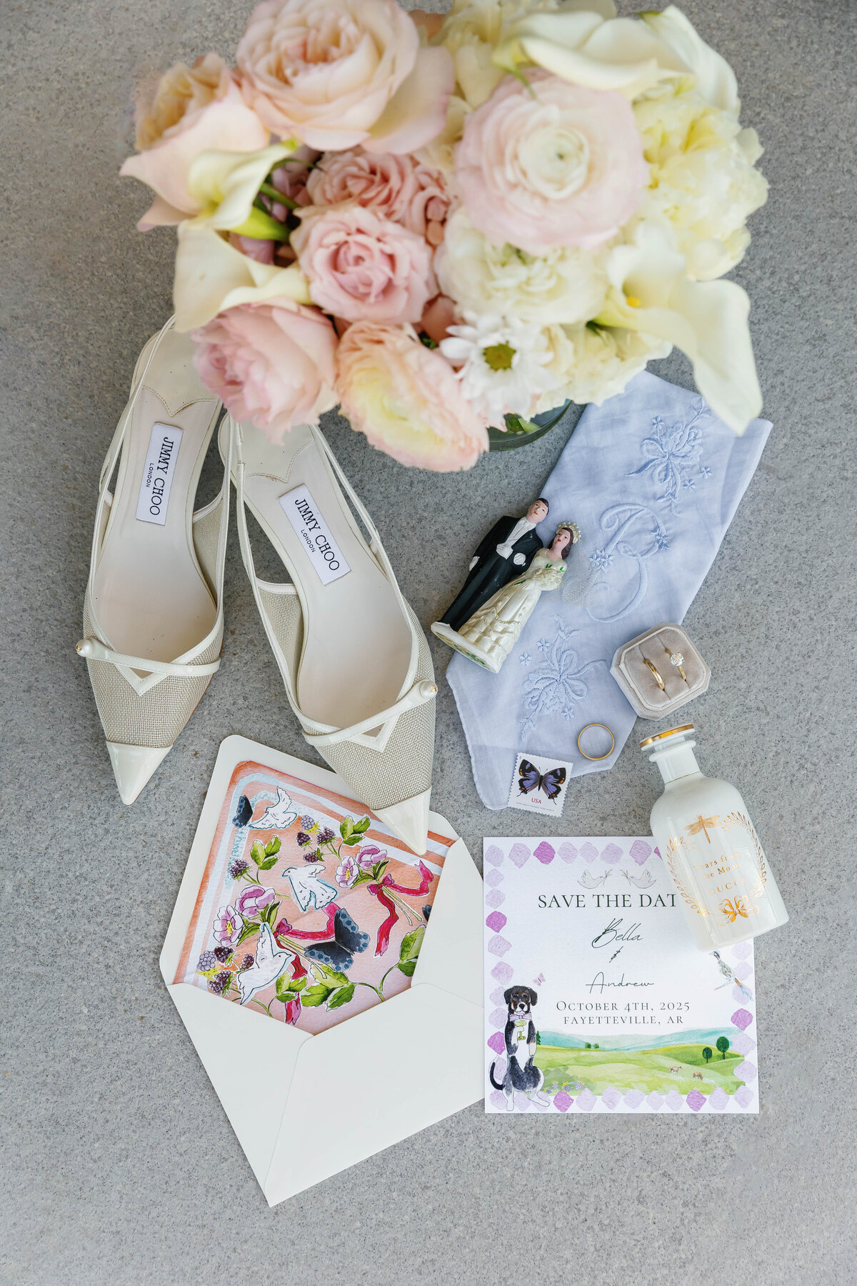 Bridal details laid out on a textured stone surface, including Jimmy Choo wedding shoes, pastel floral bouquet, embroidered blue heirloom handkerchief, vintage bride and groom figurines, engagement ring in a velvet box, wedding bands, perfume bottle, and illustrated save-the-date card. A soft, romantic flat-lay capturing meaningful wedding keepsakes.
