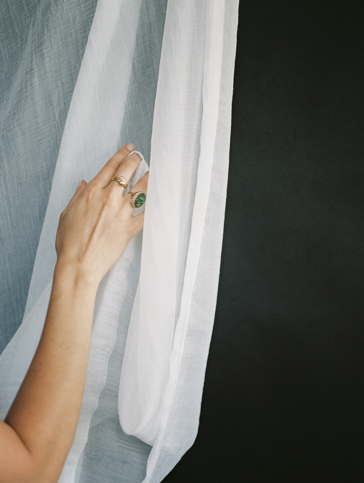 woman's hand with gold heirloom jewelry, green stone, moves white curtain fabric near black background