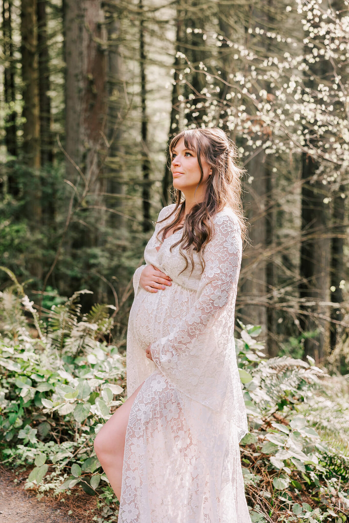 Portrait of a woman with long hair and bangs wearing white lace gown in the forest in portland, oregon. She has her hands on her belly and is looking up to the left.