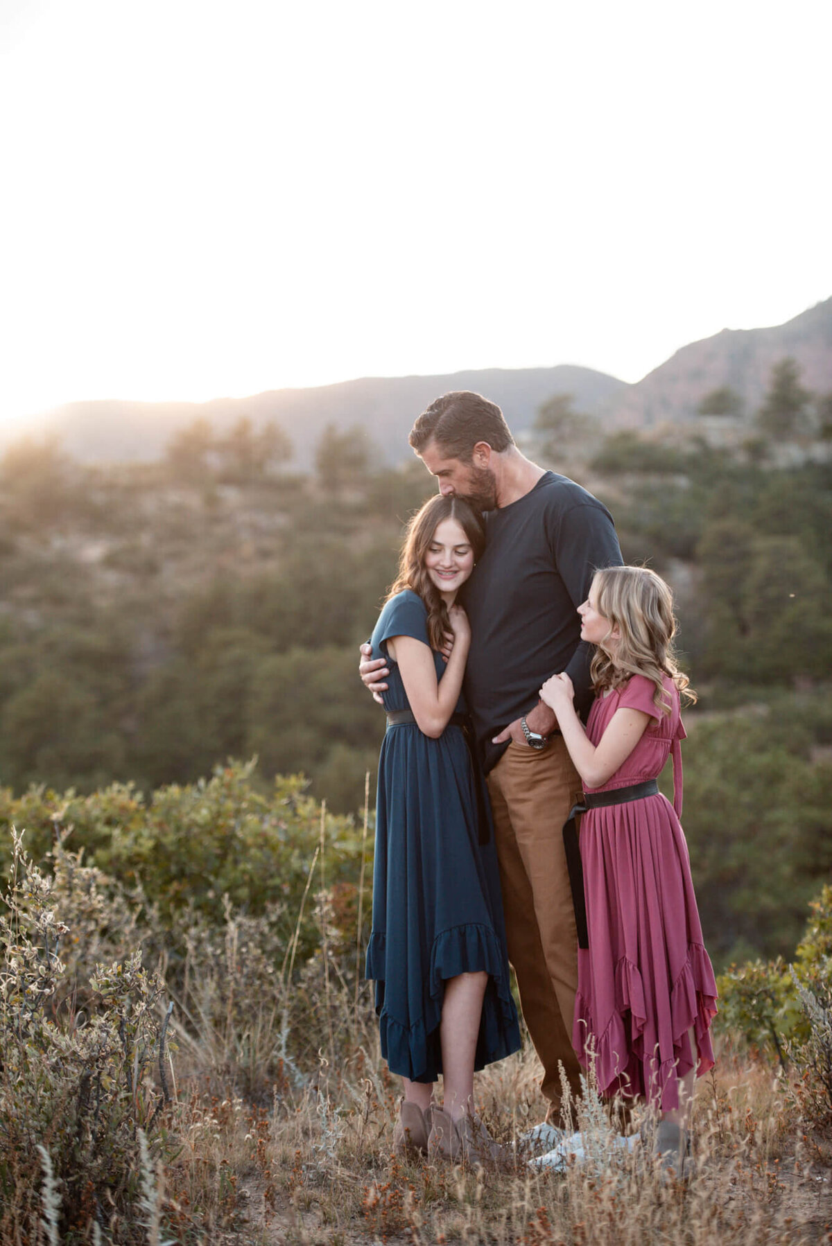 A father in a blue sweater kisses the head of his teen daughter while his other daughter hugs his arm