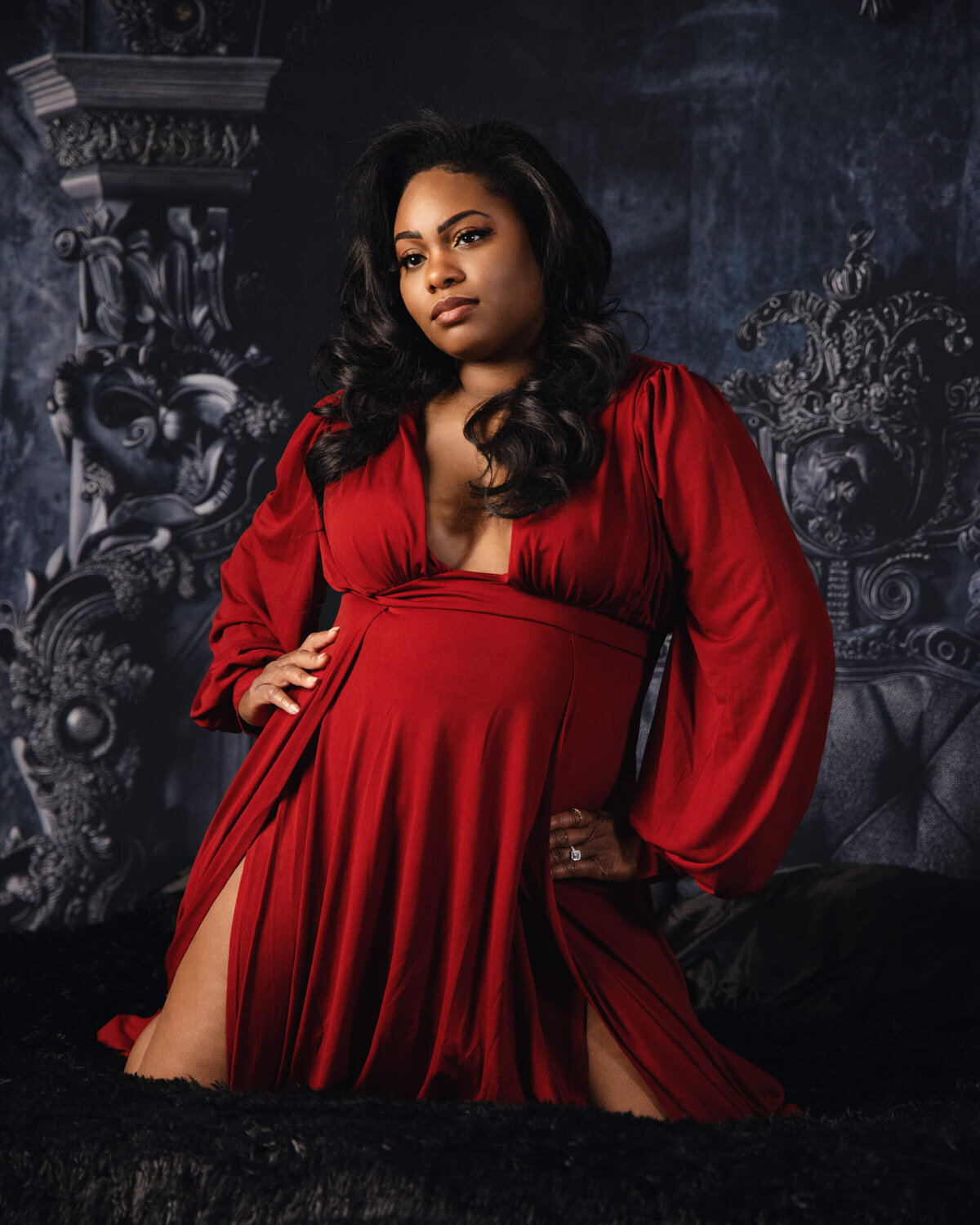 A woman wearing a flowing red dress poses confidently with one hand on her hip and the other resting on a dark surface. She is kneeling against an ornate, decorative backdrop, with an elaborately carved wooden element visible in the background.