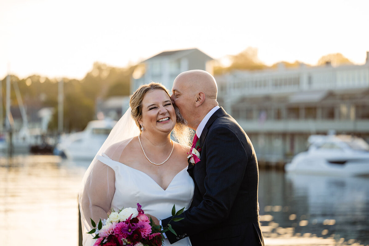 A close-up of a groom kissing a bride's cheek with waterfront buildings and boats in soft focus behind them