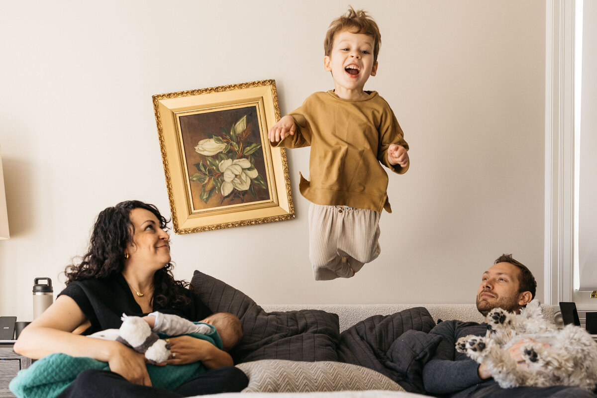 Young family are snuggling in bed with their energetic toddler son jumping. 