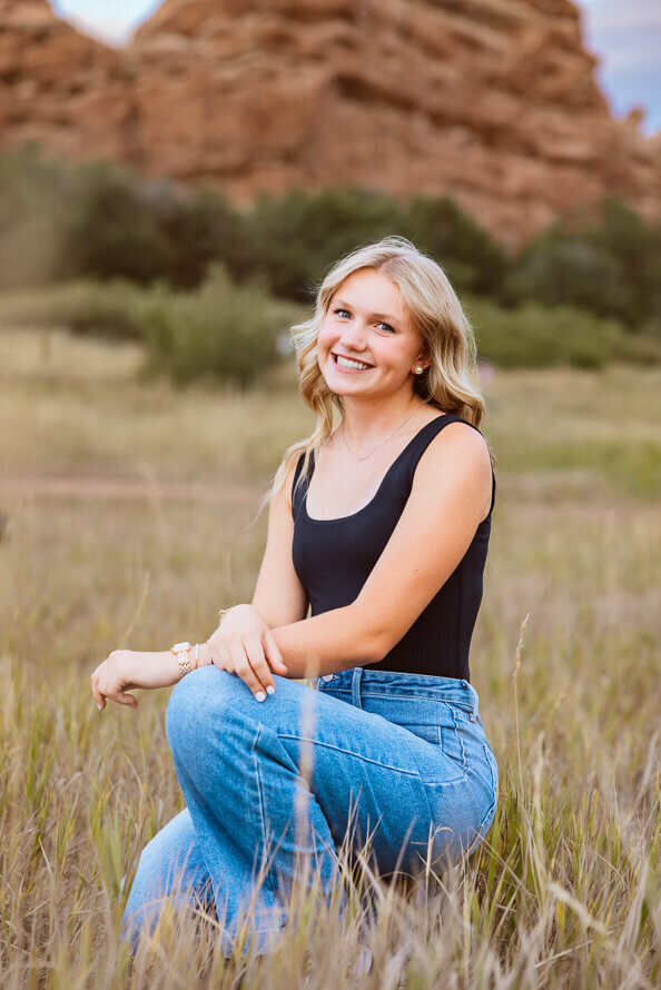 Girl squats in a grassy field for her senior session with Angela Marie Media.